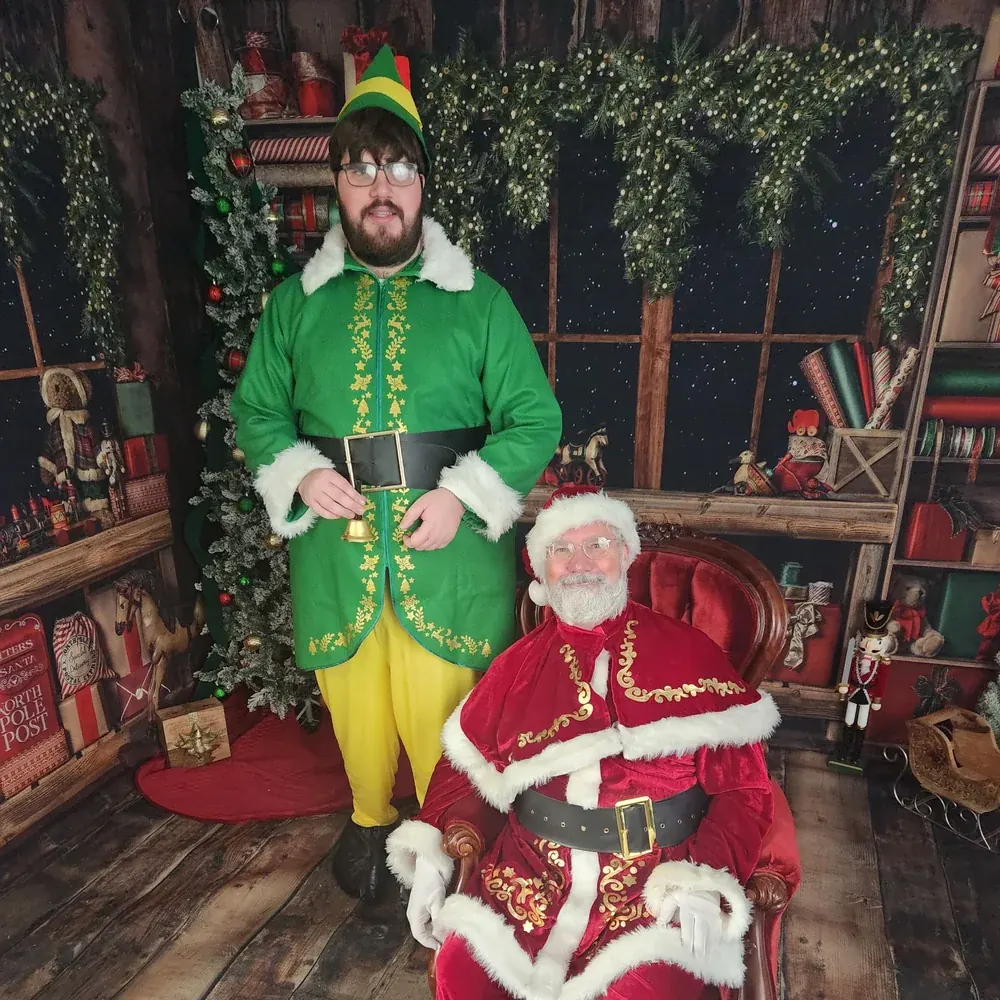 Buddy the Elf stands beside Santa Michael in a festive holiday setting.